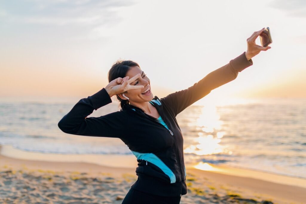young-smiling-attractive-slim-woman-doing-sport-exercises-morning-sunrise-beach-sports-wear-healthy-lifestyle-listening-music-earphones-making-selfie-photo-phone-positive-mood_285396-5538